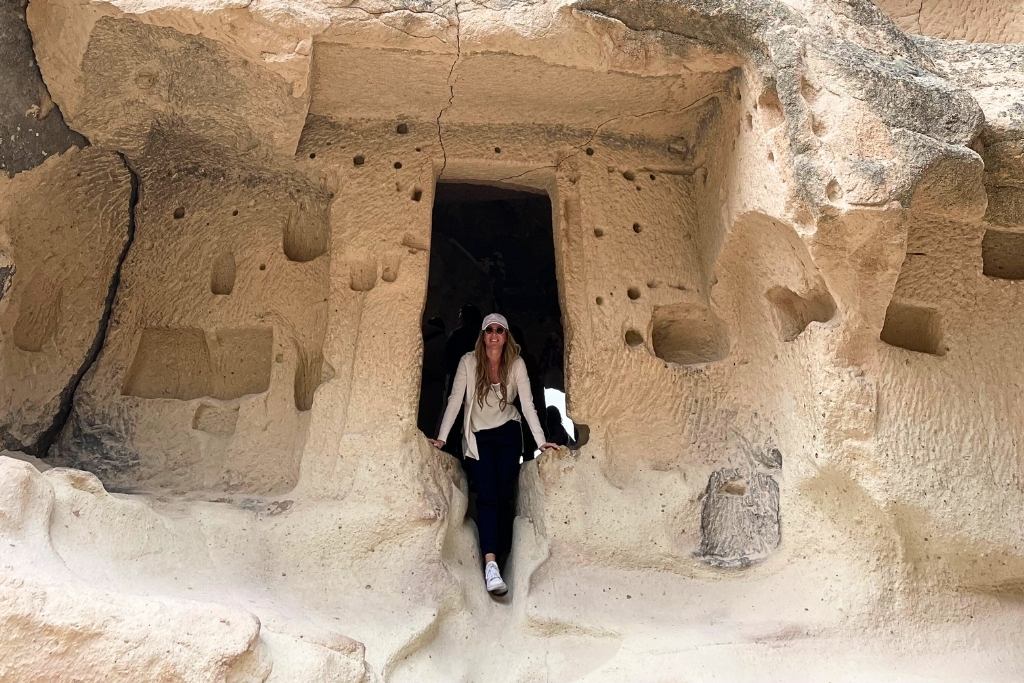 Kate stands at the entrance of an ancient carved rock dwelling in Cappadocia, surrounded by intricate stonework and natural formations. The scene highlights the region's unique and historic architecture.