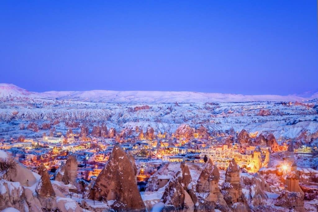 A breathtaking winter view of Cappadocia with snow-covered fairy chimneys and rock formations illuminated by the warm glow of village lights. The vibrant blue and purple hues of the twilight sky enhance the magical atmosphere of the scene.