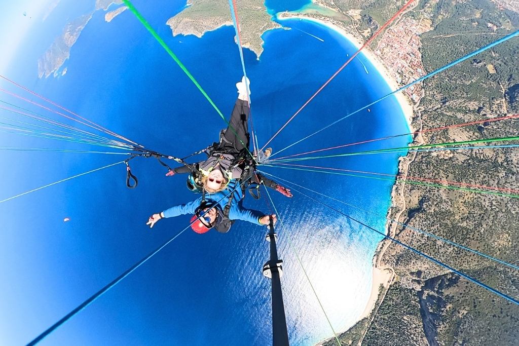 Kate paraglides high above the turquoise waters and sandy beaches of Ölüdeniz, Turkey, with a wide view of the coastline and surrounding mountains. The colorful parachute cords frame the exhilarating scene as she smiles mid-flight.