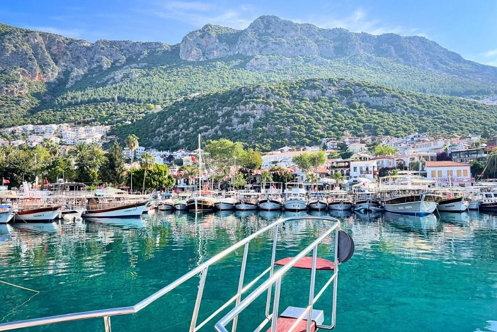 A picturesque harbor in Kaş, Turkey, with boats docked on clear turquoise water and colorful buildings nestled against a lush green hillside. The scene is framed by towering mountains under a bright blue sky.