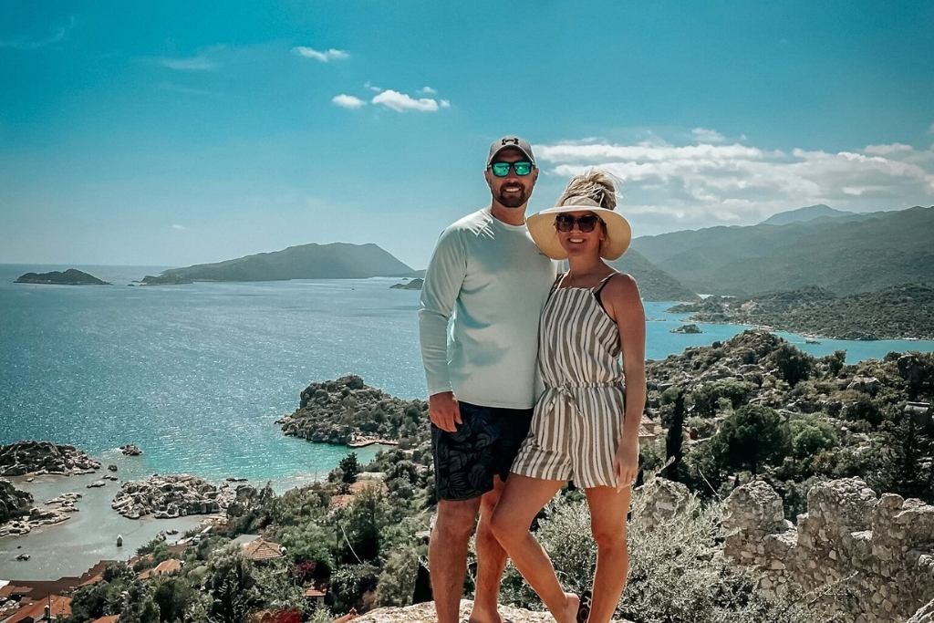 Kate and her husband pose together on a hillside overlooking the stunning turquoise waters and rocky islands of the Turkish coastline. The woman wears a striped romper and wide-brimmed hat, while the man is dressed casually in a cap and sunglasses. The backdrop features lush greenery and a peaceful, sunlit seascape.