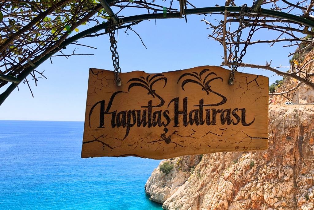 A rustic wooden sign reading 'Kaputaş Hatırası' hangs from a metal archway, with a clear view of the deep blue Mediterranean Sea and rocky cliffs in the background. 