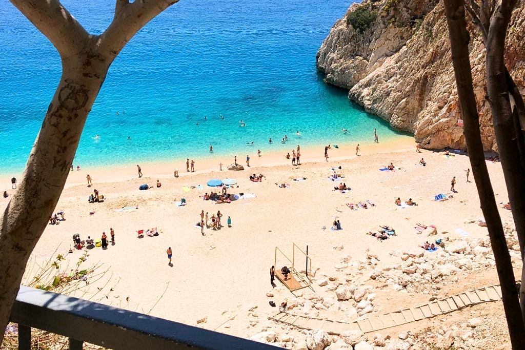 An overhead view of Kaputaş Beach in Turkey, showcasing golden sand, turquoise waters, and people sunbathing and swimming along the shoreline. The rocky cliffs frame the serene beach, while a wooden pathway leads down to the sand.