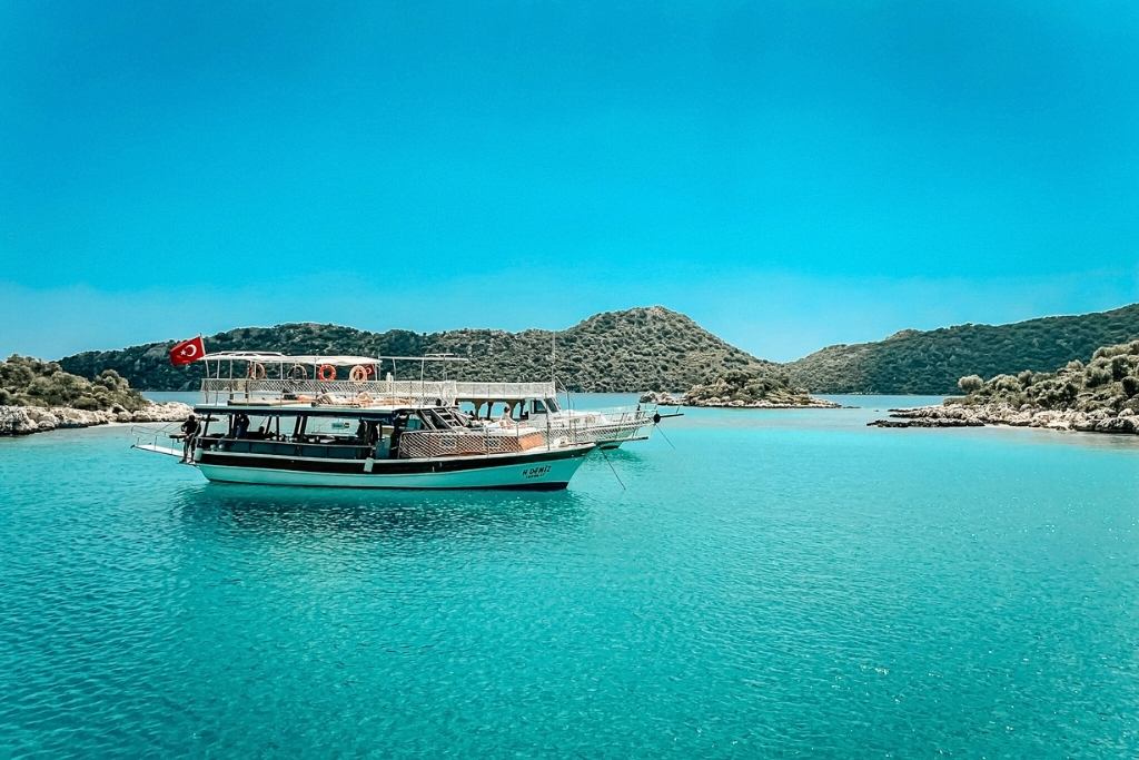 A traditional wooden boat with a Turkish flag floats on the crystal-clear turquoise waters of the Turquoise Coast in Turkey. The serene scene is surrounded by lush green hills and a bright blue sky.