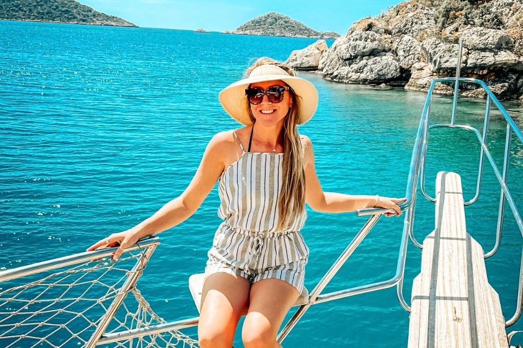 Kate, in a striped romper, sunglasses, and a wide-brimmed hat, sits on the railing of a boat, surrounded by the turquoise waters of the Turkish coast. The backdrop features rocky islands and lush hills under a bright, sunny sky.