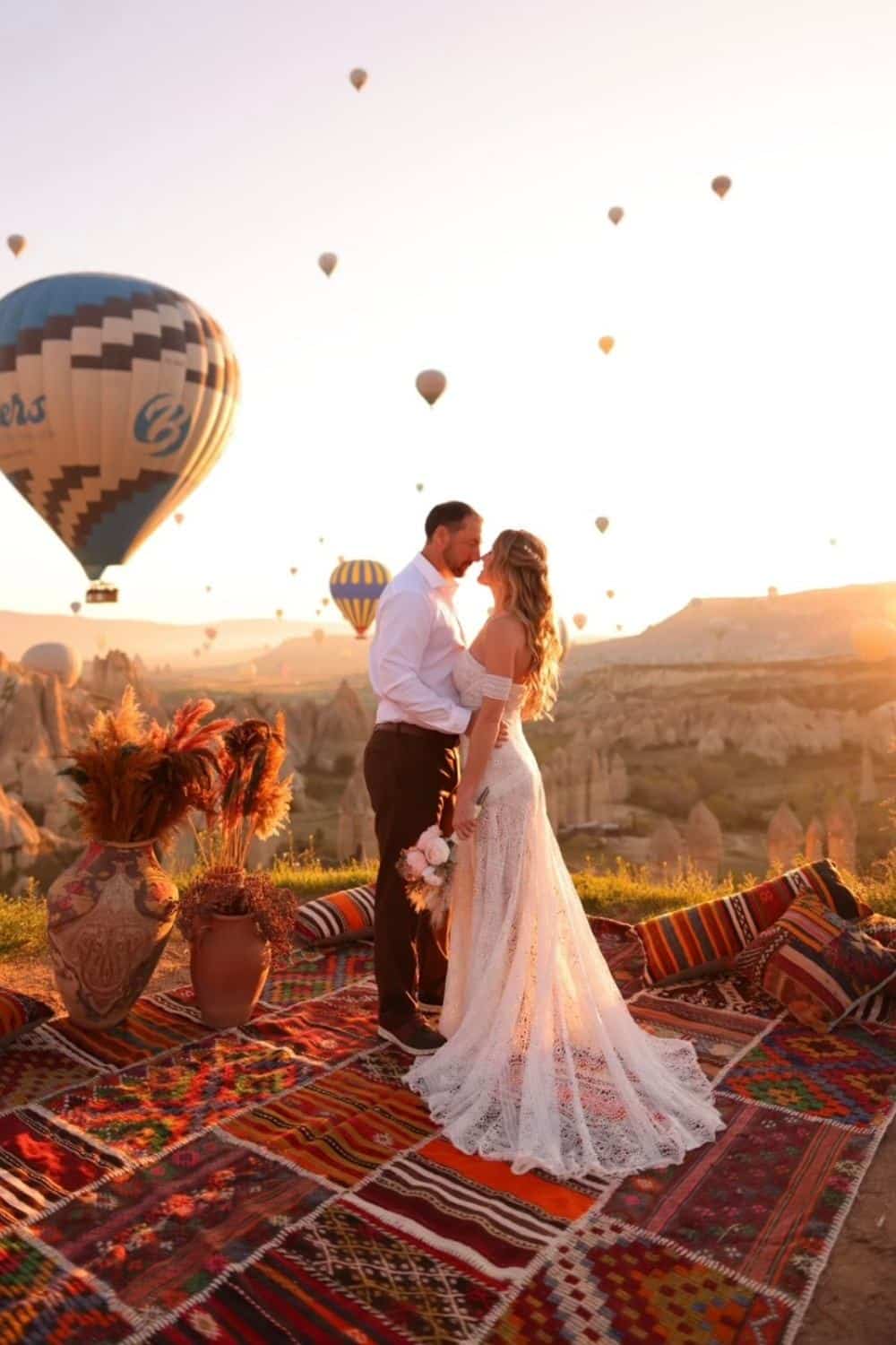 Kate from Kate's Crossing Blog and her husband sharing a romantic moment on a colorful Turkish rug overlooking Cappadocia's iconic landscape at sunrise. Hot air balloons float in the sky, creating a dreamy backdrop, while Kate holds a bouquet and wears a flowing lace dress, and her husband is dressed in a white shirt and dark pants.