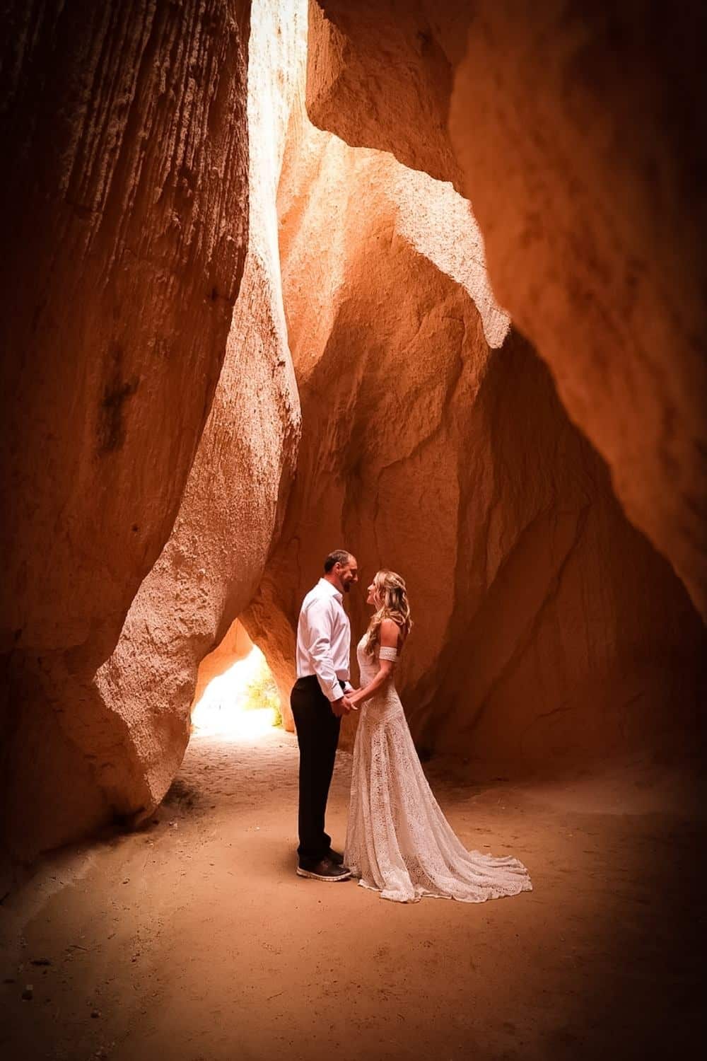 Kate from Kate's Crossing Blog and her husband standing together in a warm, glowing sandstone canyon in Turkey. The couple is holding hands, with Kate wearing a flowing lace gown and her husband dressed in a white shirt and dark trousers. The light streaming in from the canyon opening highlights the romantic atmosphere.