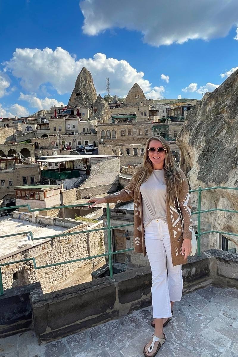 Kate from Kate's Crossing Blog standing on a terrace in Cappadocia with traditional stone buildings and fairy chimneys in the background. She is wearing a patterned brown cardigan, white pants, and sunglasses, smiling against the bright blue sky with fluffy clouds. The unique architecture and rock formations highlight the beauty of the Cappadocian landscape.