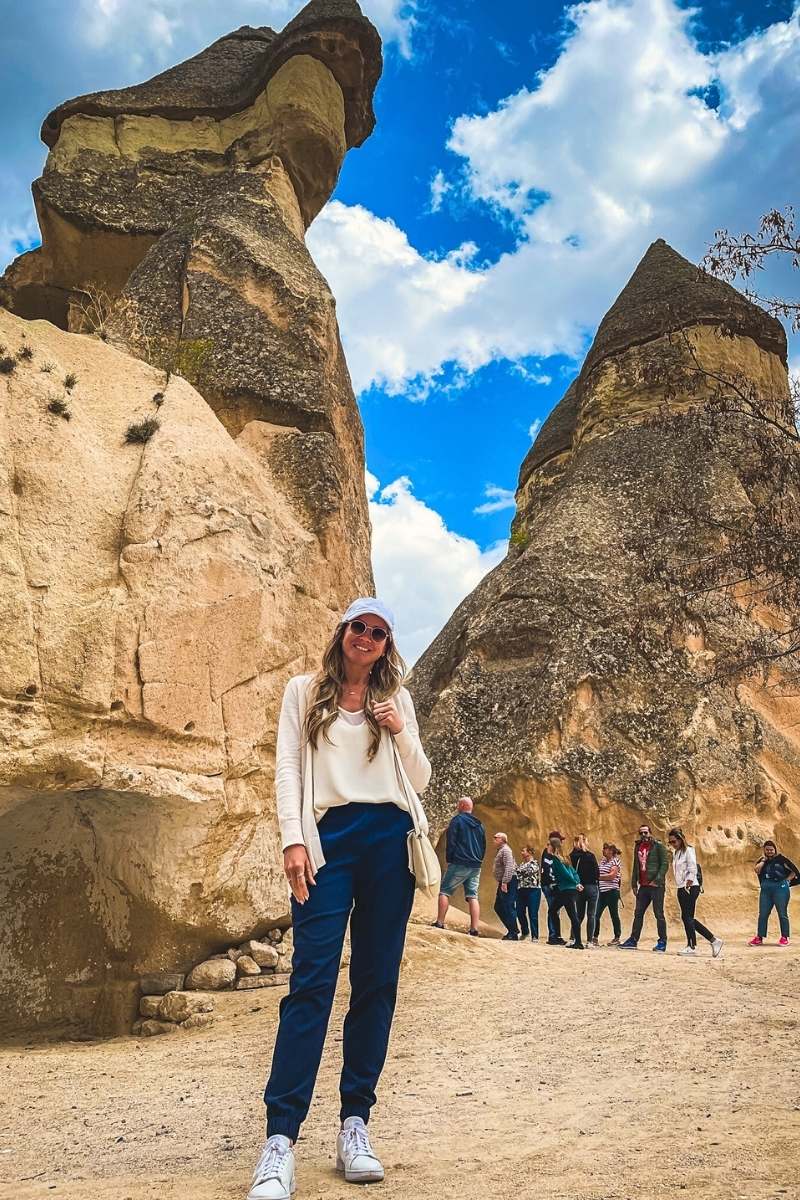 Kate from Kate's Crossing Blog standing in front of the distinctive fairy chimneys in Cappadocia, Turkey. She is wearing a white top, a beige cardigan, navy pants, sneakers, and a white cap, smiling under the bright blue sky with scattered clouds. The unique rock formations and the small group of tourists in the background capture the essence of this famous geological site.