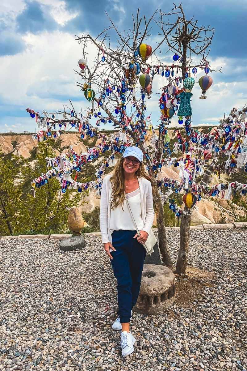 Kate from Kate's Crossing Blog standing in front of a decorative evil eye tree in Cappadocia, Turkey. The tree is adorned with colorful Nazar charms, ribbons, and miniature hot air balloon ornaments, set against a backdrop of rocky hills and cloudy skies. She is wearing a white top, beige cardigan, navy pants, sneakers, and a white cap, smiling as she enjoys the unique cultural display.