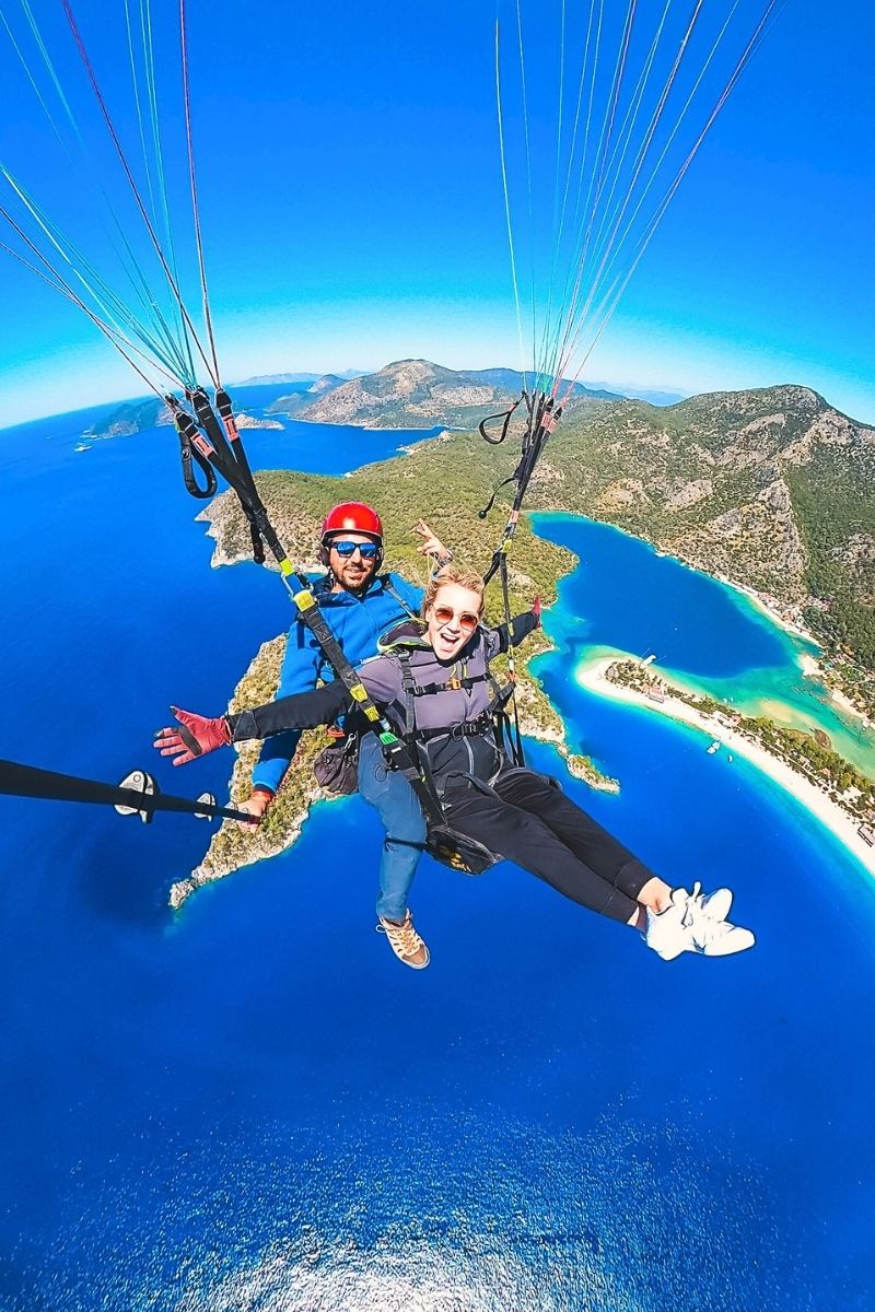 Kate from Kate's Crossing Blog paragliding high above the stunning blue coastline and green hills of Ölüdeniz, Turkey. She is smiling widely, wearing sunglasses and harnessed with her instructor, as they glide over the vibrant turquoise waters and sandy beaches below. The breathtaking aerial view captures the thrill and beauty of the experience.