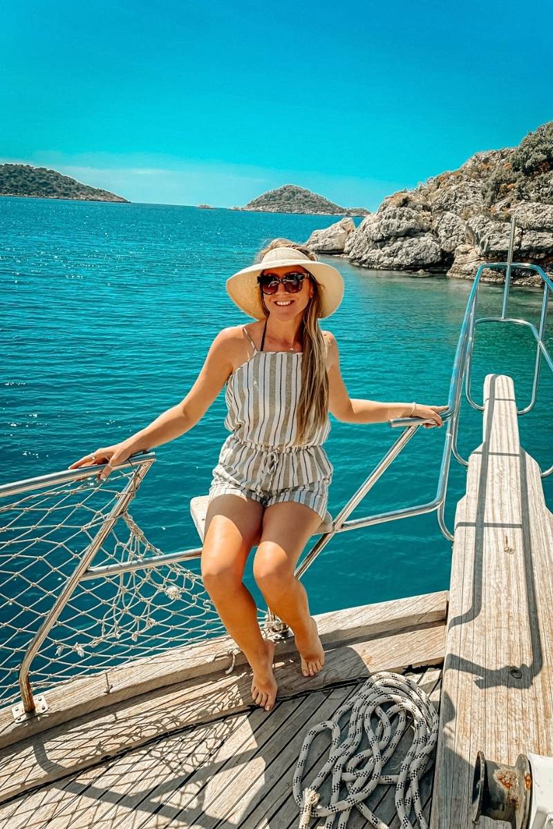 Kate from Kate's Crossing Blog relaxing on the deck of a boat, surrounded by the clear turquoise waters of Turkey's coastline. She is wearing a striped romper, a wide-brimmed straw hat, and sunglasses, smiling under the bright sun. The serene sea and rocky islands in the background create a peaceful and picturesque scene.