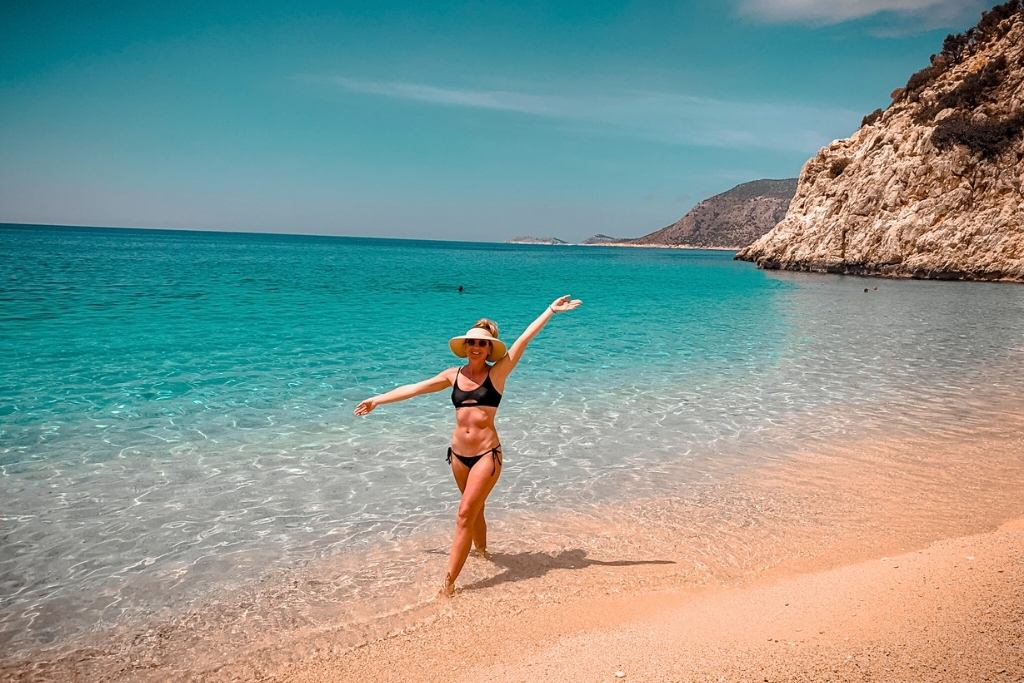 Kate from Kate's Crossing Blog enjoying a sunny day on a pristine beach in Turkey, standing at the water's edge with her arms outstretched. She is wearing a black bikini and a wide-brimmed straw hat, with turquoise waves gently lapping at the shore. The rocky cliffs and clear blue sky create a serene and inviting seaside backdrop.