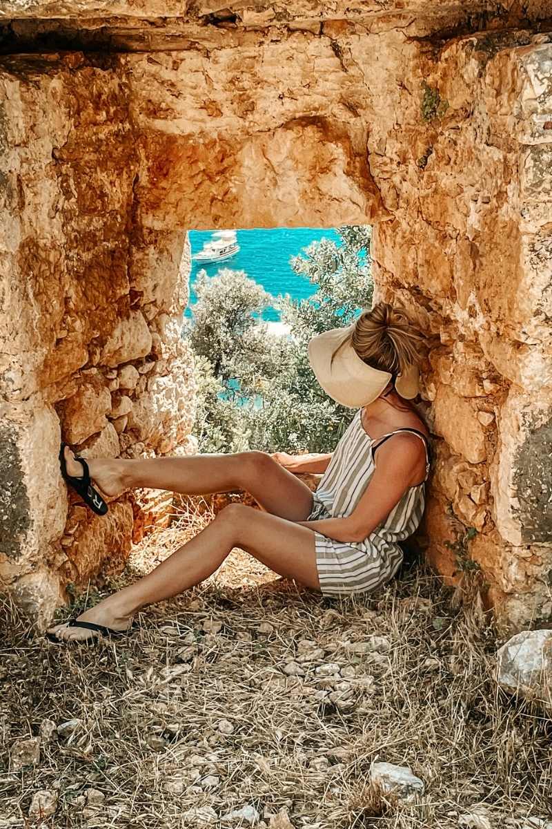 Kate from Kate's Crossing Blog sitting in a stone archway of a historic ruin in Turkey, gazing out at the turquoise waters below. She is wearing a striped romper, a wide-brimmed visor hat, and flip-flops, relaxed against the rustic stone walls. The scenic view of the sea and boats through the arch adds to the charm of the tranquil moment.