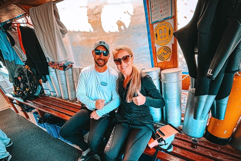 Kate from Kate's Crossing Blog and her husband sitting on a bench aboard a dive boat, surrounded by scuba tanks and wetsuits. Both are wearing casual athletic clothing, sunglasses, and smiling with thumbs up, ready for an underwater adventure. The lively atmosphere and gear emphasize the excitement of a diving trip in Turkey.