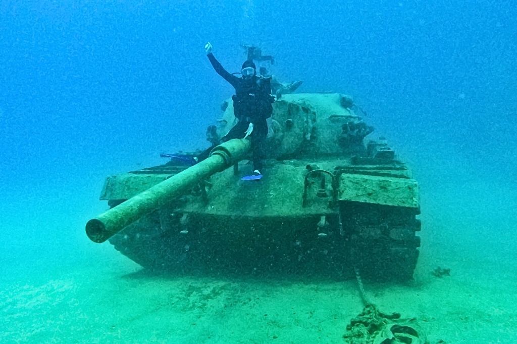 Kate from Kate's Crossing Blog, posing underwater on the barrel of a sunken tank in Turkey. The tank is covered in marine growth and rests on the sandy ocean floor, surrounded by the clear blue depths. The scene captures a unique underwater adventure with an intriguing historical artifact.