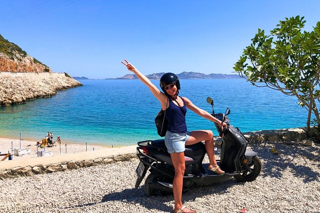 Kate from Kate's Crossing Blog posing on a scooter near a picturesque beach in Turkey, with turquoise water and rocky cliffs in the background. She is wearing a black helmet, navy top, denim shorts, and sandals, raising one arm in excitement. The clear blue sky and vibrant sea emphasize the fun and adventurous vibe of the coastal setting.