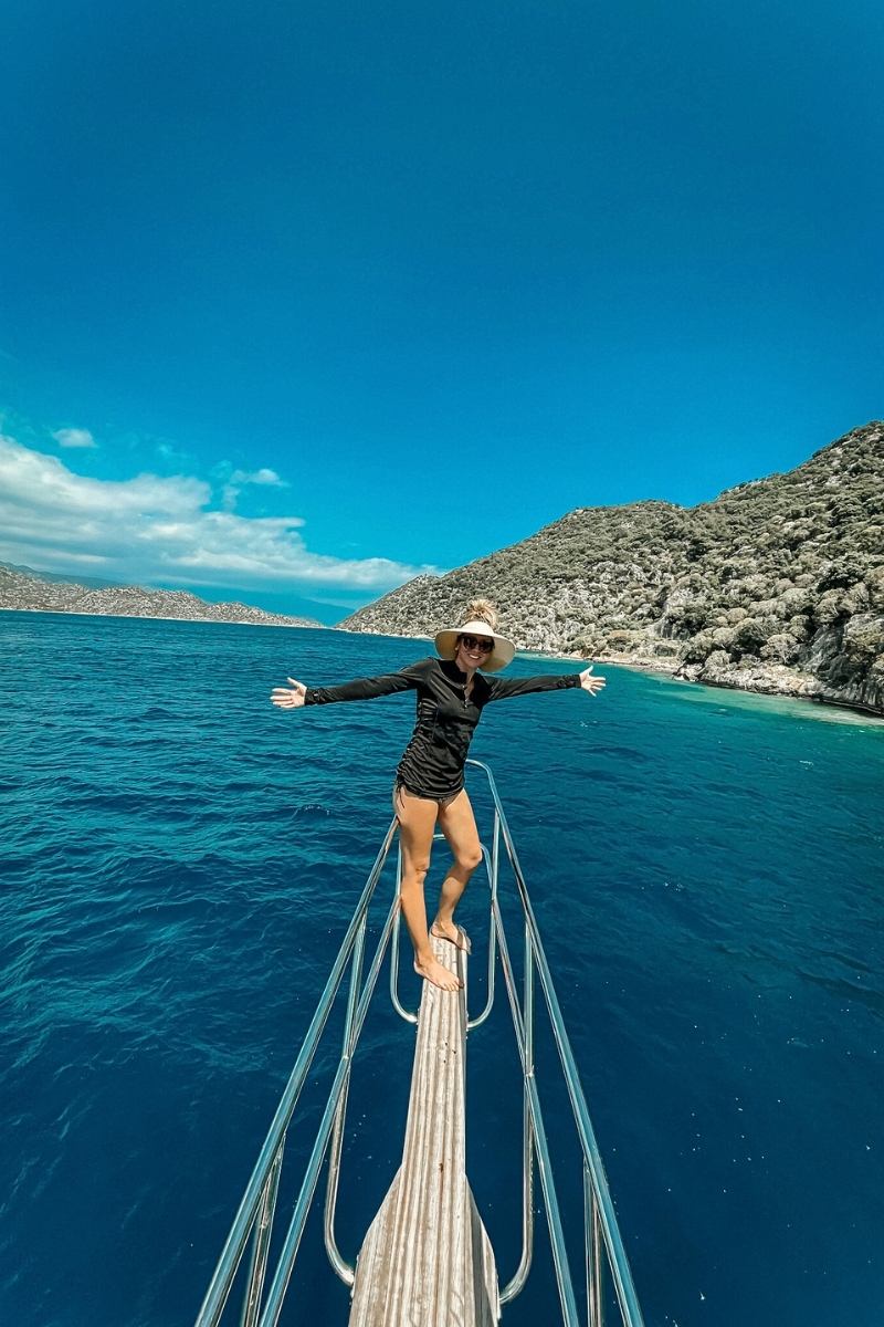 Kate from Kate's Crossing Blog standing at the bow of a boat with arms outstretched, surrounded by the deep turquoise waters of Turkey’s coastline. She is wearing a black cover-up, a wide-brimmed hat, and sandals, smiling under a sunny blue sky. The lush green hills and rocky shoreline in the background complete the scenic and adventurous moment.