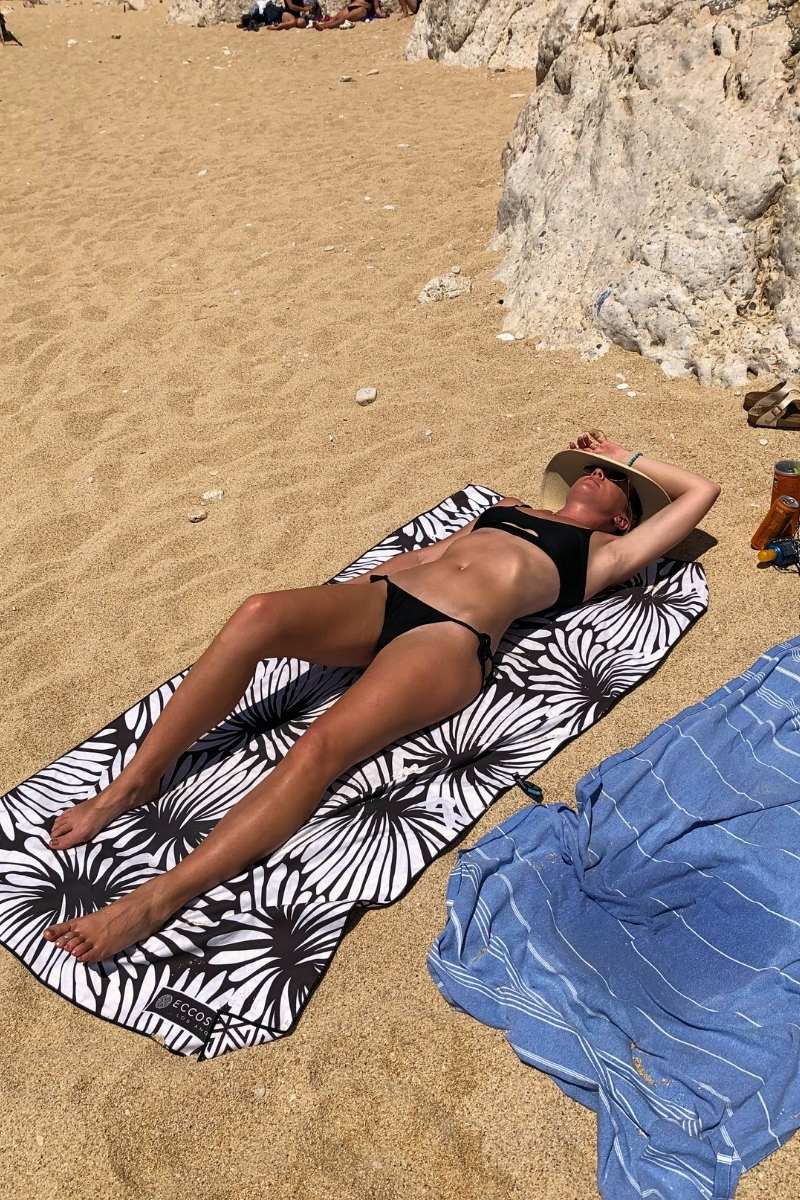 Kate from Kate's Crossing Blog sunbathing on a sandy beach in Turkey, lying on a patterned black-and-white beach towel. She is wearing a black bikini and a wide-brimmed hat, relaxing near a rocky backdrop. A blue towel and sunscreen bottle are nearby, highlighting the tranquil and sunny seaside setting.