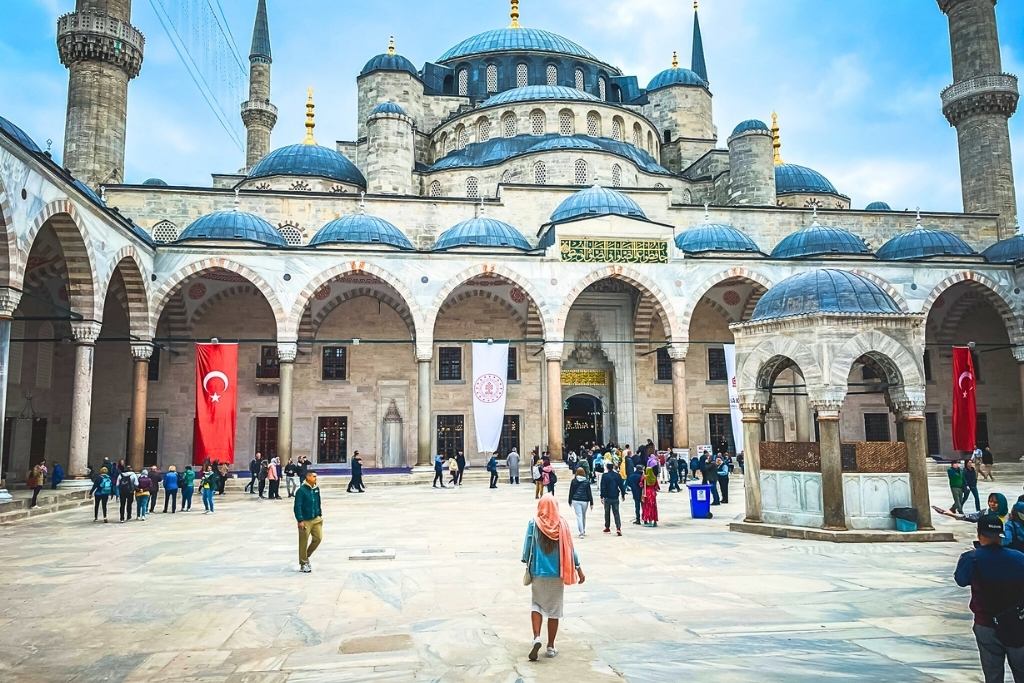 Kate from Kate's Crossing Blog walking through the courtyard of the Blue Mosque in Istanbul, Turkey. She is wearing a pink scarf and modest attire, surrounded by the mosque's grand arches, domes, and intricate architecture. The Turkish flag and a marble fountain add to the vibrant and historic atmosphere of the iconic site.