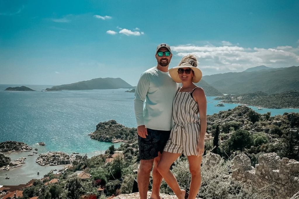 Kate from Kate's Crossing Blog and her husband posing on a rocky overlook with a stunning view of the turquoise sea and distant hills in Turkey. Kate is wearing a striped romper and a wide-brimmed hat, while her husband is in a light shirt and shorts, both smiling under a bright blue sky.