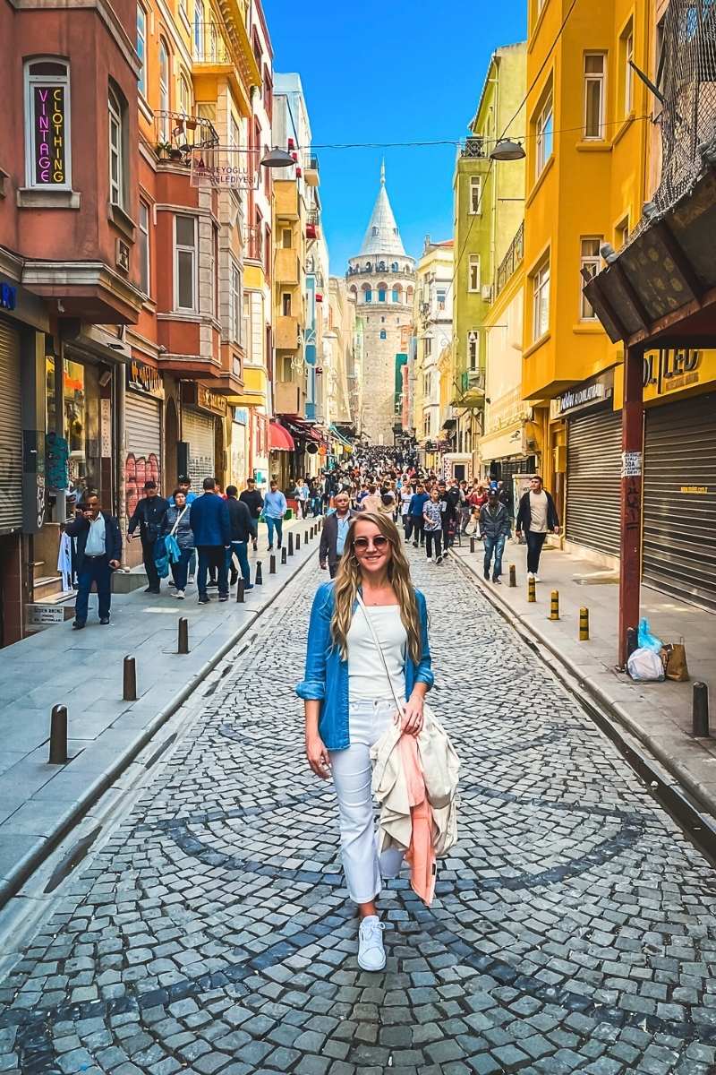 Kate from Kate's Crossing Blog standing on a cobblestone street in Istanbul with the iconic Galata Tower visible in the background. She is dressed casually in white pants, a blue denim shirt, and sneakers, holding a light jacket, with colorful buildings and bustling crowds surrounding her.