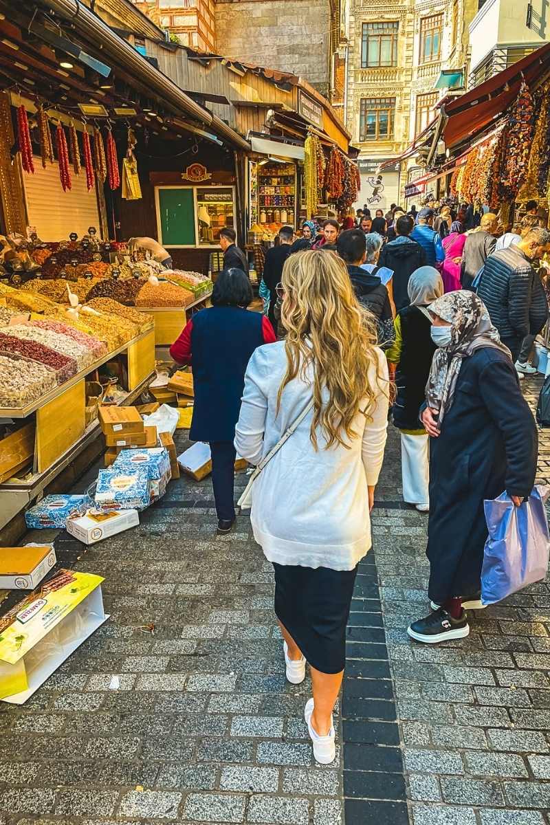 Kate from Kate's Crossing Blog walking through a bustling Turkish market filled with colorful displays of spices, nuts, and dried goods. She is wearing a white cardigan, a black skirt, and sneakers, blending into the lively crowd of shoppers under the vibrant canopy of market stalls.