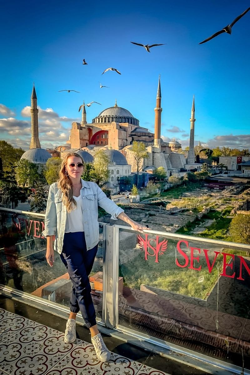 Kate from Kate's Crossing Blog standing on a rooftop terrace with the Hagia Sophia in Istanbul as the stunning backdrop. She is wearing a denim jacket, white top, navy pants, and sneakers, with seagulls soaring in the bright blue sky above.