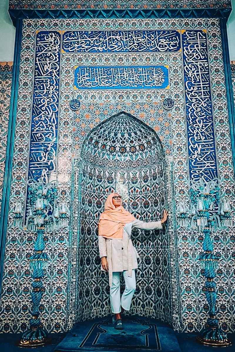 Kate from Kate's Crossing Blog standing in front of an intricate and colorful tiled mihrab inside a mosque in Turkey. She is dressed modestly, wearing a light pink scarf and beige coat, looking up at the detailed Islamic calligraphy and geometric patterns that adorn the wall.