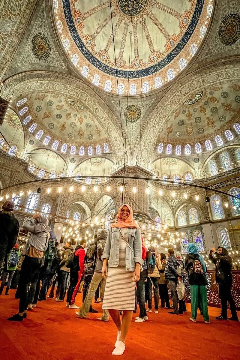 Kate from Kate's Crossing Blog standing inside the magnificent Blue Mosque in Istanbul, surrounded by intricate domes adorned with Islamic calligraphy and stunning stained glass windows. She is wearing a pink scarf, a striped dress, and a denim jacket, smiling as soft lighting from chandeliers illuminates the mosque's grandeur.