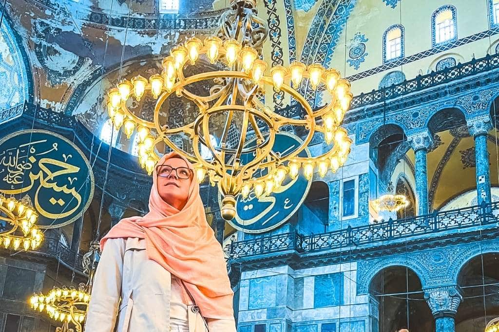 Kate from Kate's Crossing Blog standing inside the Hagia Sophia in Istanbul, illuminated by the glow of ornate chandeliers hanging from the ceiling. She is wearing a light pink scarf and a beige coat, surrounded by the historic mosque's stunning arches, intricate Islamic calligraphy, and rich architectural details.