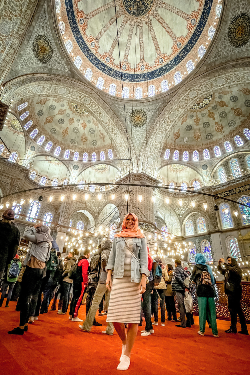 Kate in a peach hijab and denim jacket stands barefoot on the red carpet inside the Blue Mosque in Istanbul, surrounded by visitors, with the mosque’s grand domes, intricate tilework, and glowing chandeliers above her.