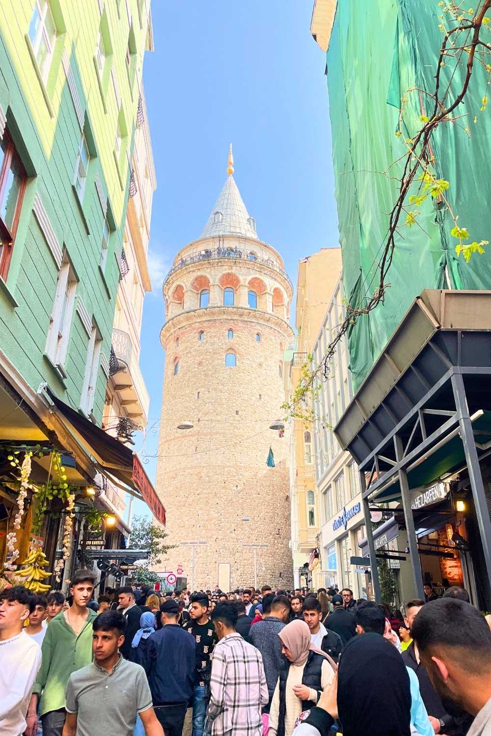 A lively street in Istanbul filled with people walking among colorful buildings, cafés, and market stalls, leading up to the iconic Galata Tower standing tall against a clear blue sky.