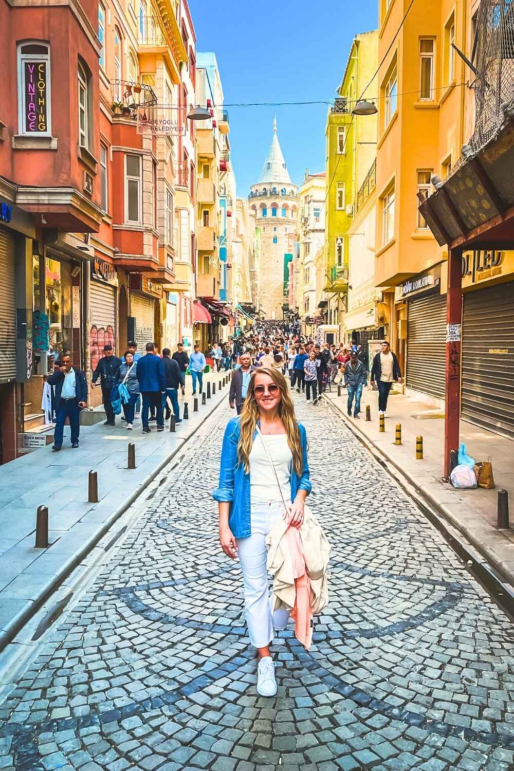 A smiling Kate in a denim jacket and white pants walks along a cobblestone street in Istanbul, with the colorful buildings of Beyoğlu and the iconic Galata Tower rising in the background amid bustling crowds.