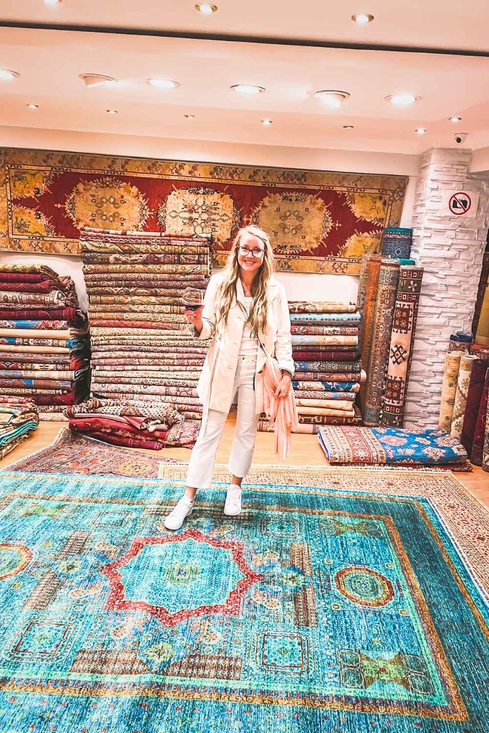 in a beige coat and white pants, holds a glass of red wine while standing in a Turkish carpet shop surrounded by stacks of intricately woven rugs in rich colors and patterns.