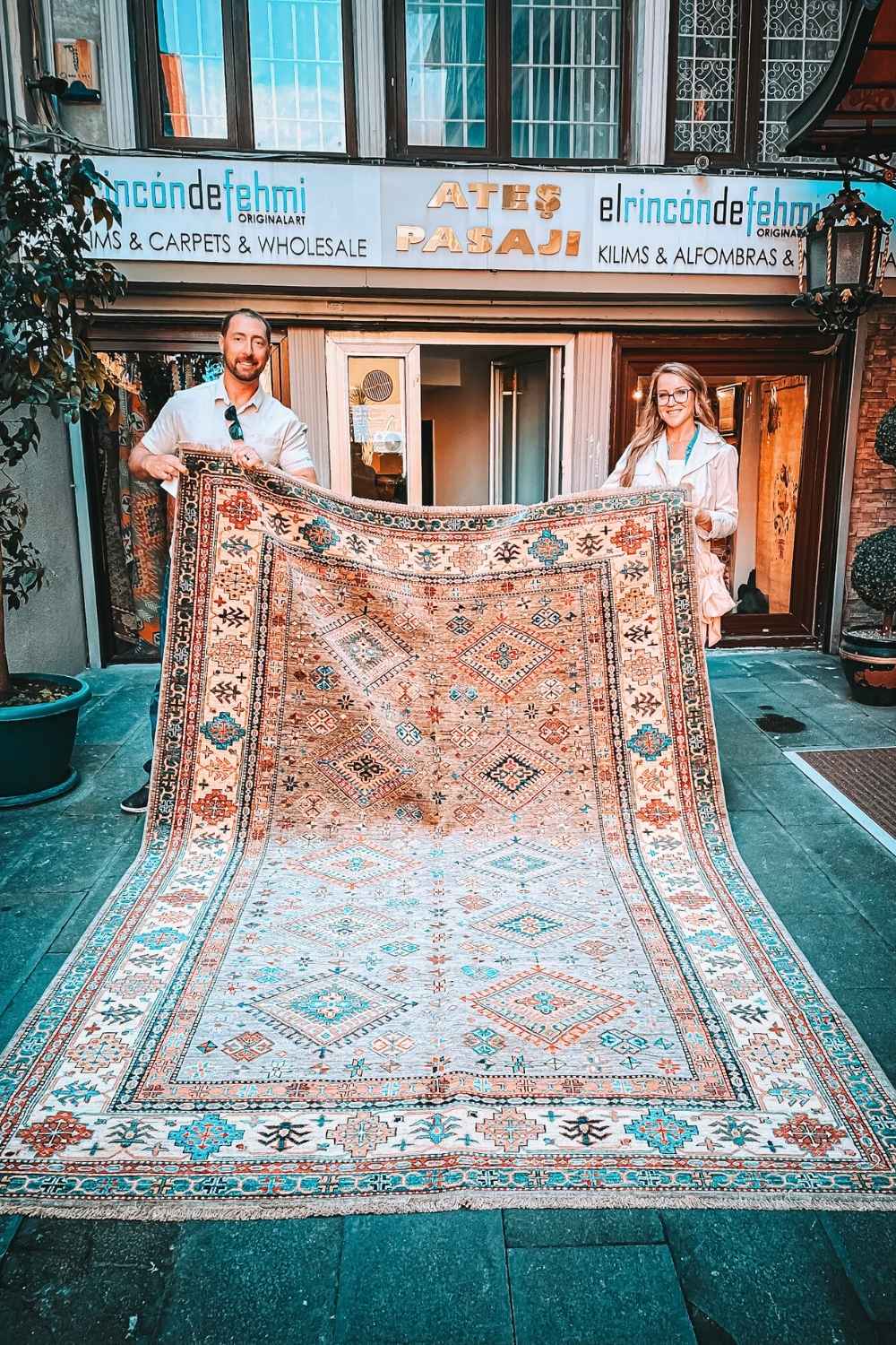 A smiling Kate and her husband hold up a beautifully patterned Turkish rug outside a carpet shop in Istanbul, with intricate geometric designs and vibrant colors.