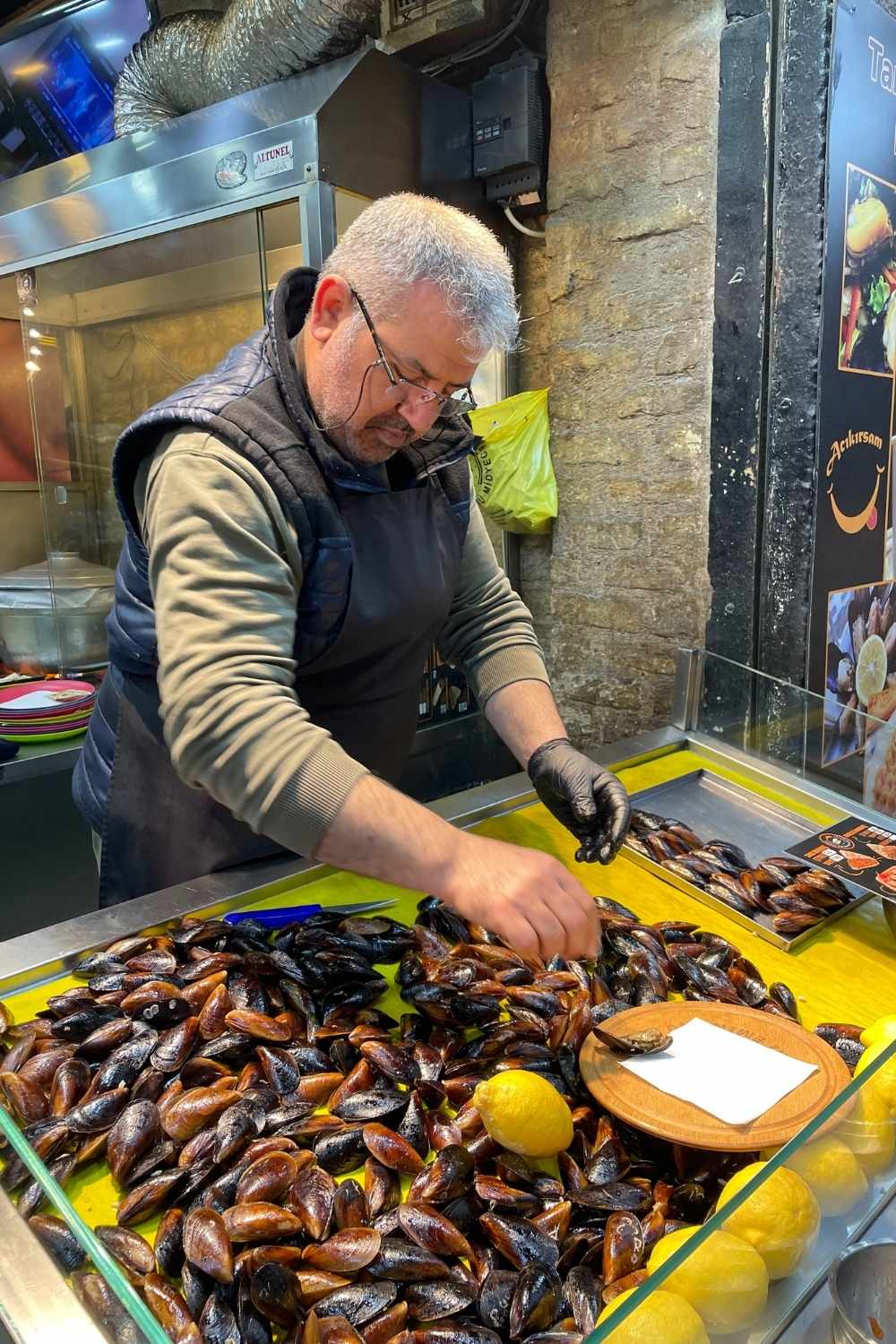 A street food vendor in Istanbul, wearing gloves and an apron, carefully prepares stuffed mussels (midye dolma) at his stall, surrounded by fresh mussels and lemons on a glass counter.