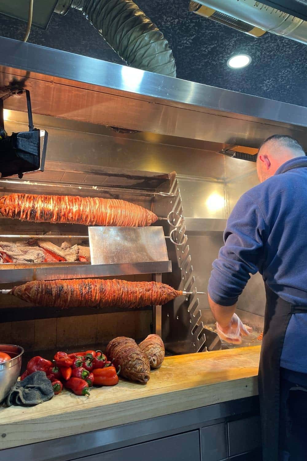 A street food vendor in Istanbul prepares kokoreç, a traditional Turkish dish made from seasoned lamb intestines, as large skewers of the delicacy roast over an open flame beside fresh red peppers on a wooden counter.