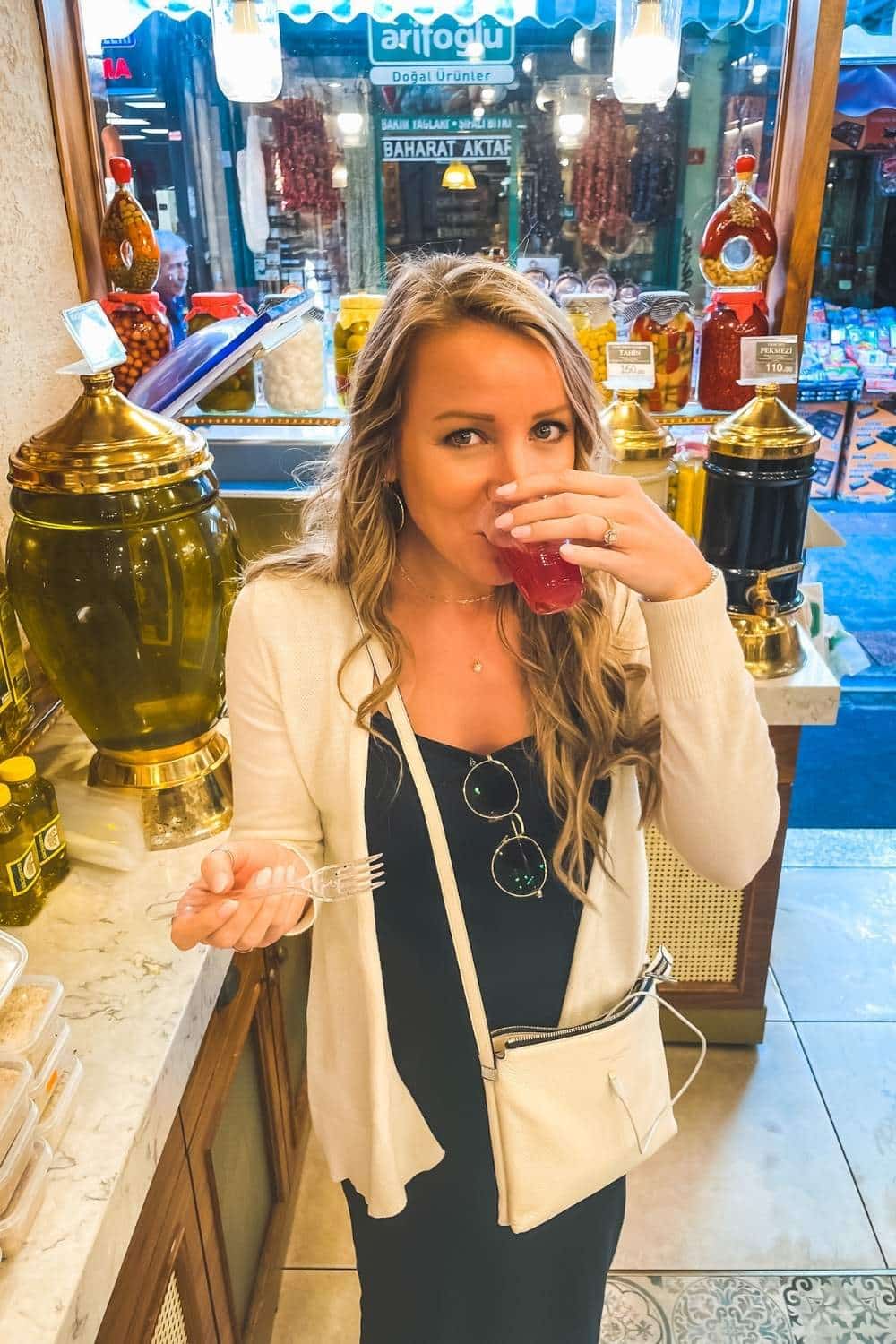 Kate, in a black dress and beige cardigan, sips on a glass of red pickle juice inside a shop, surrounded by jars of spices, oils, and traditional products, with a bustling market visible through the window behind her.