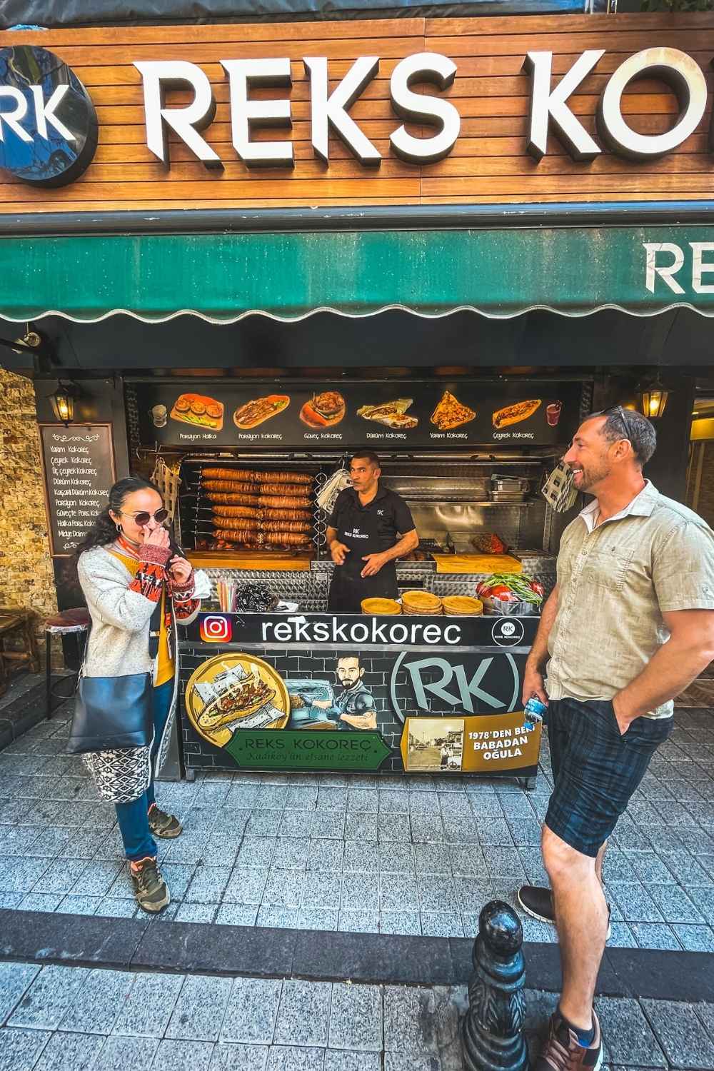 A man and woman stand in front of a kokoreç street food stall in Istanbul, chatting with the vendor as skewers of the traditional grilled dish roast behind him, under a sign reading "Reks Kokoreç."