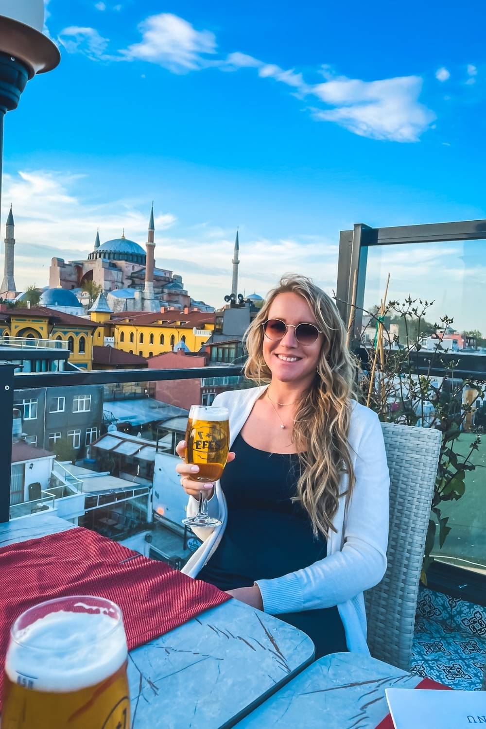 Kate in sunglasses and a white cardigan enjoys a cold glass of Efes beer on a rooftop terrace in Istanbul, with the iconic Hagia Sophia and its towering minarets in the background under a clear blue sky.