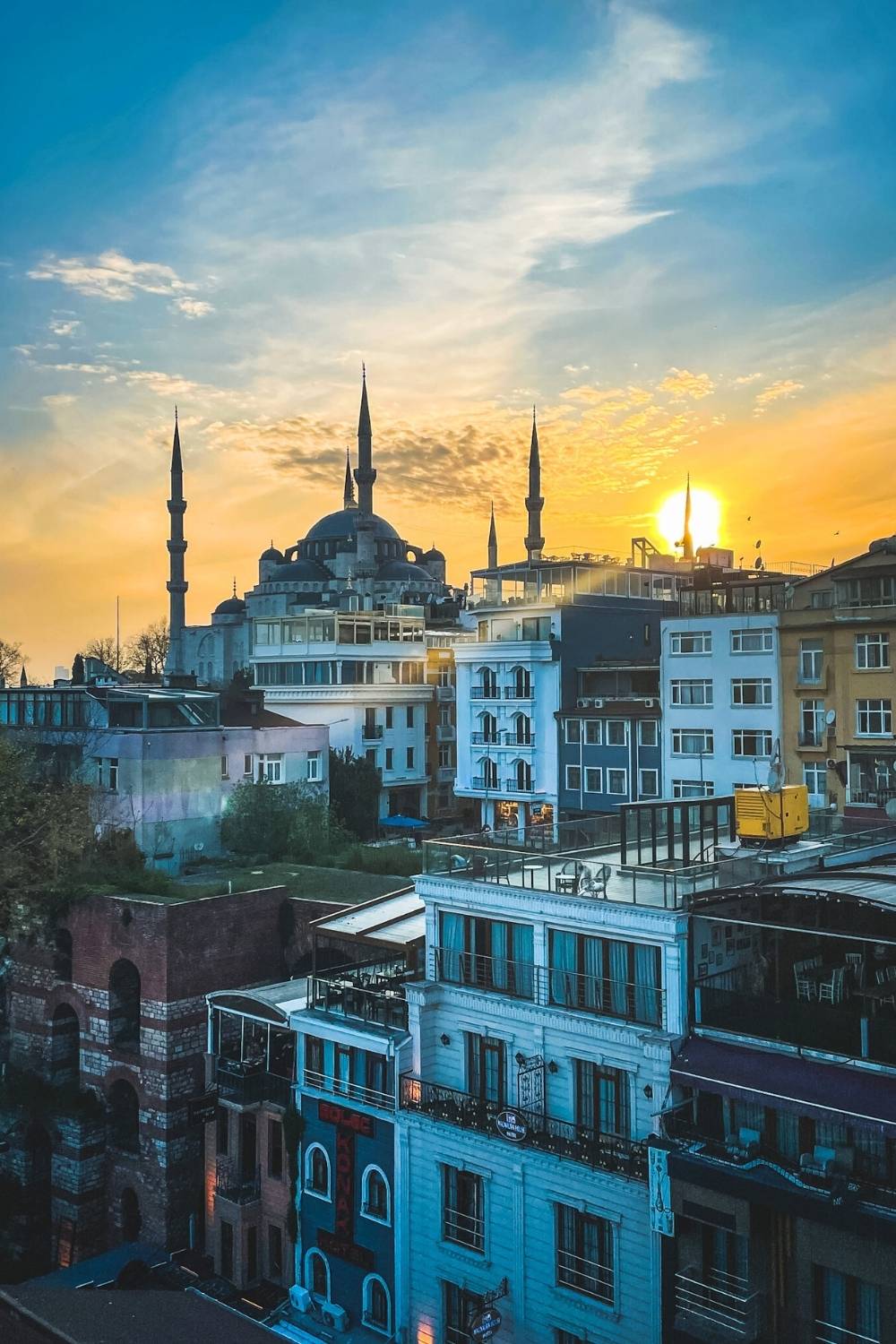 A breathtaking sunset over Istanbul, with the silhouette of the Blue Mosque and its towering minarets glowing against a golden sky, surrounded by colorful buildings and rooftop terraces.