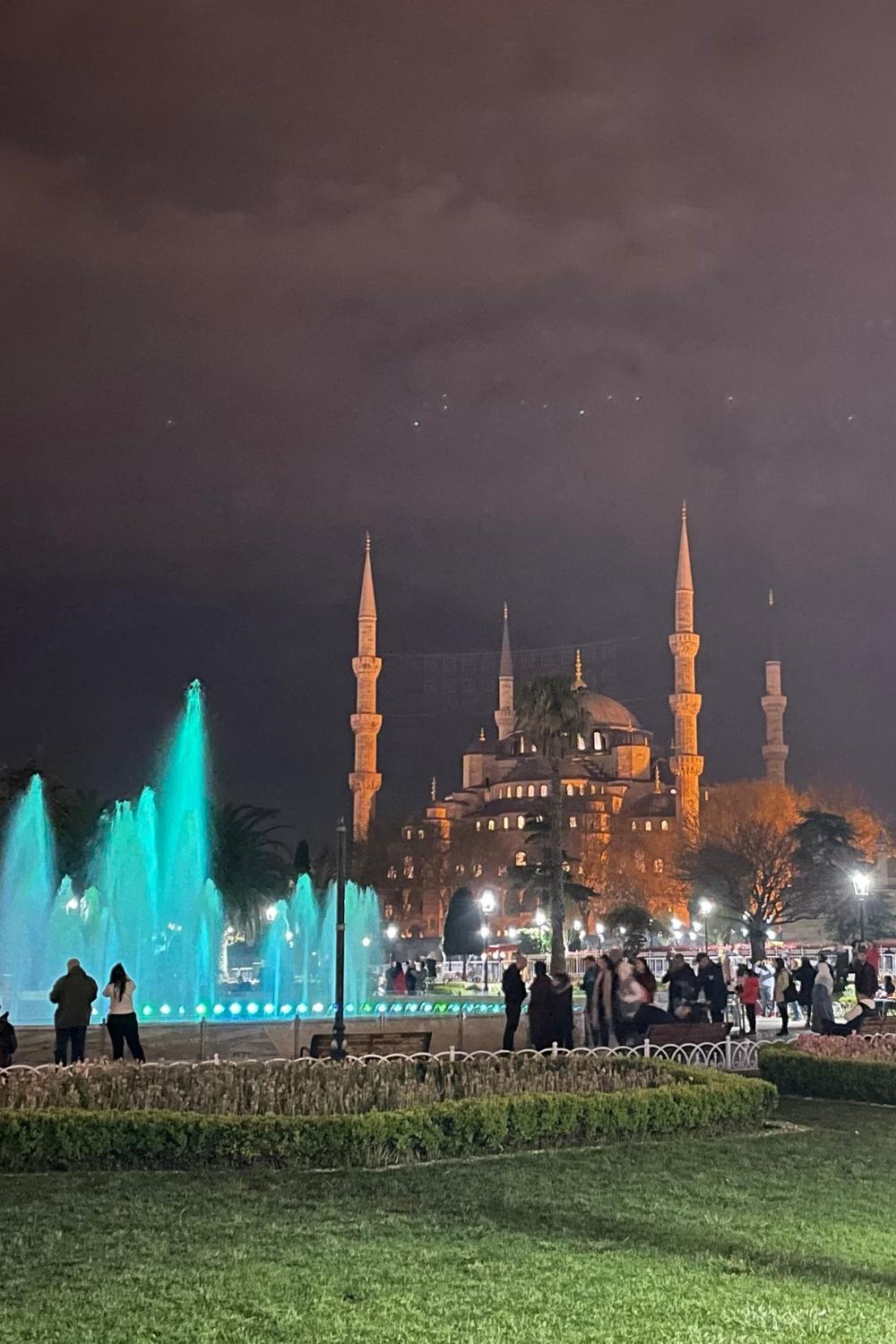 The illuminated Blue Mosque in Istanbul stands against the night sky, with glowing turquoise fountains in the foreground and people strolling through the lively park.