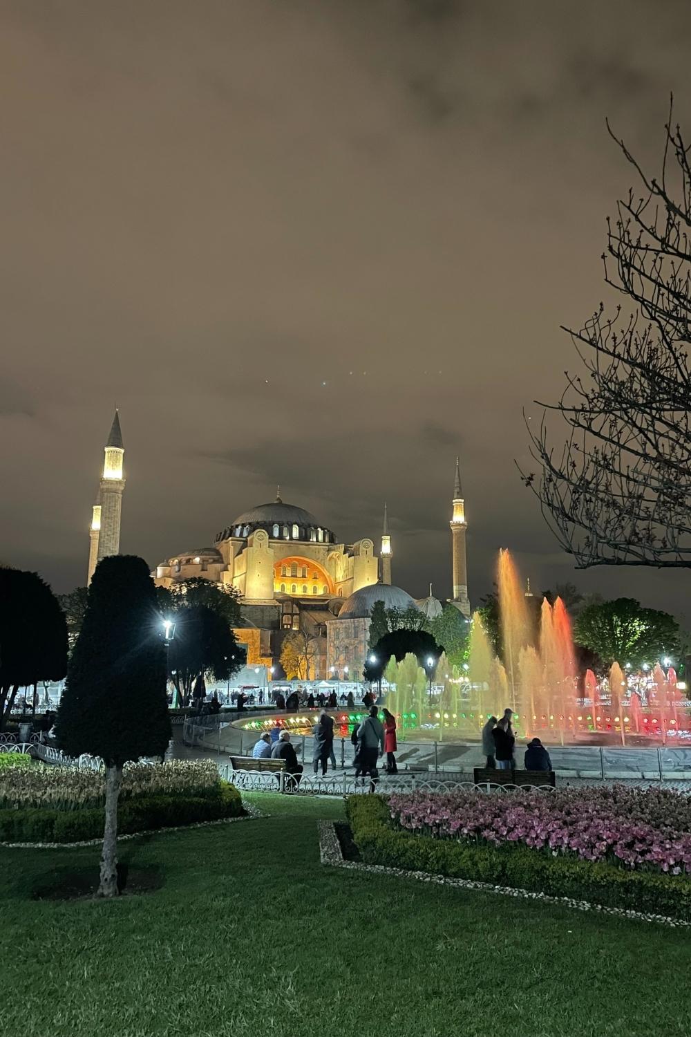 The illuminated Hagia Sophia stands majestically against a cloudy night sky in Istanbul, with colorful fountains glowing in the foreground and visitors strolling through the scenic park.