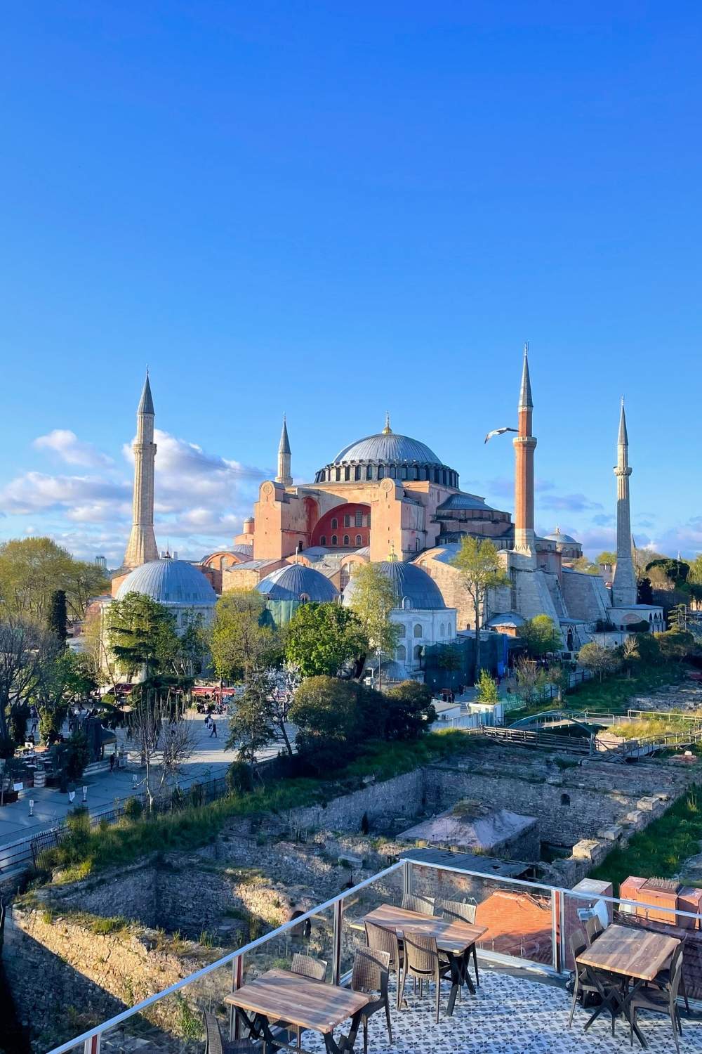 The iconic domes and towering minarets of Hagia Sophia stand against a vibrant blue sky. An outdoor terrace with wooden tables overlooks the historic site.