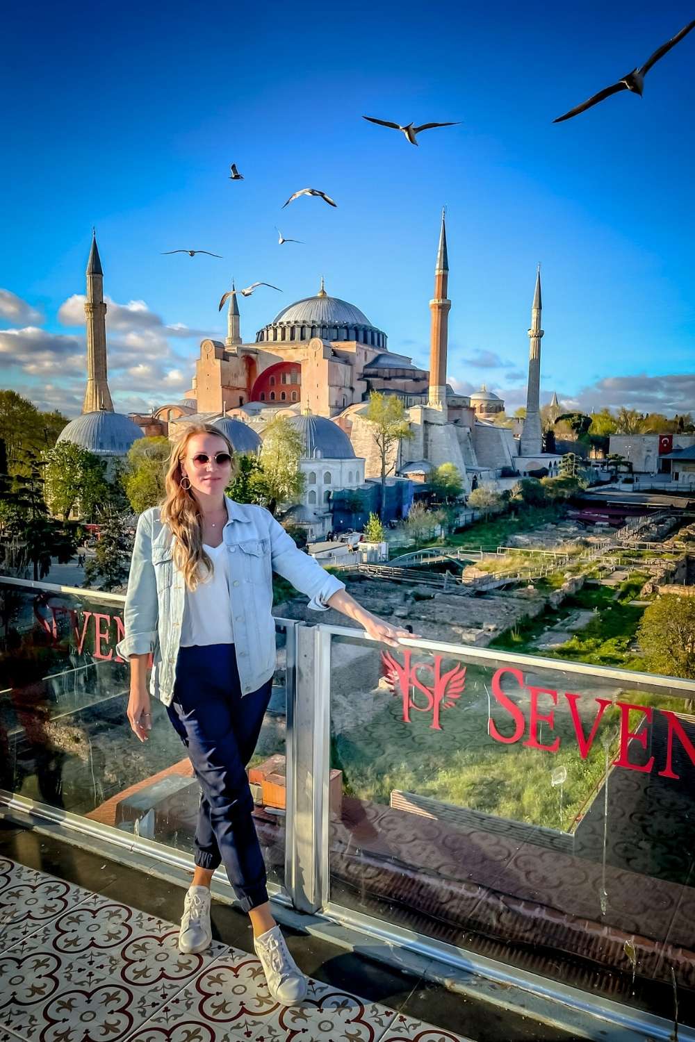 Kate in a denim jacket stands on a glass-walled rooftop overlooking the Hagia Sophia, with seagulls flying overhead. The domes and minarets stand tall under a bright blue sky.