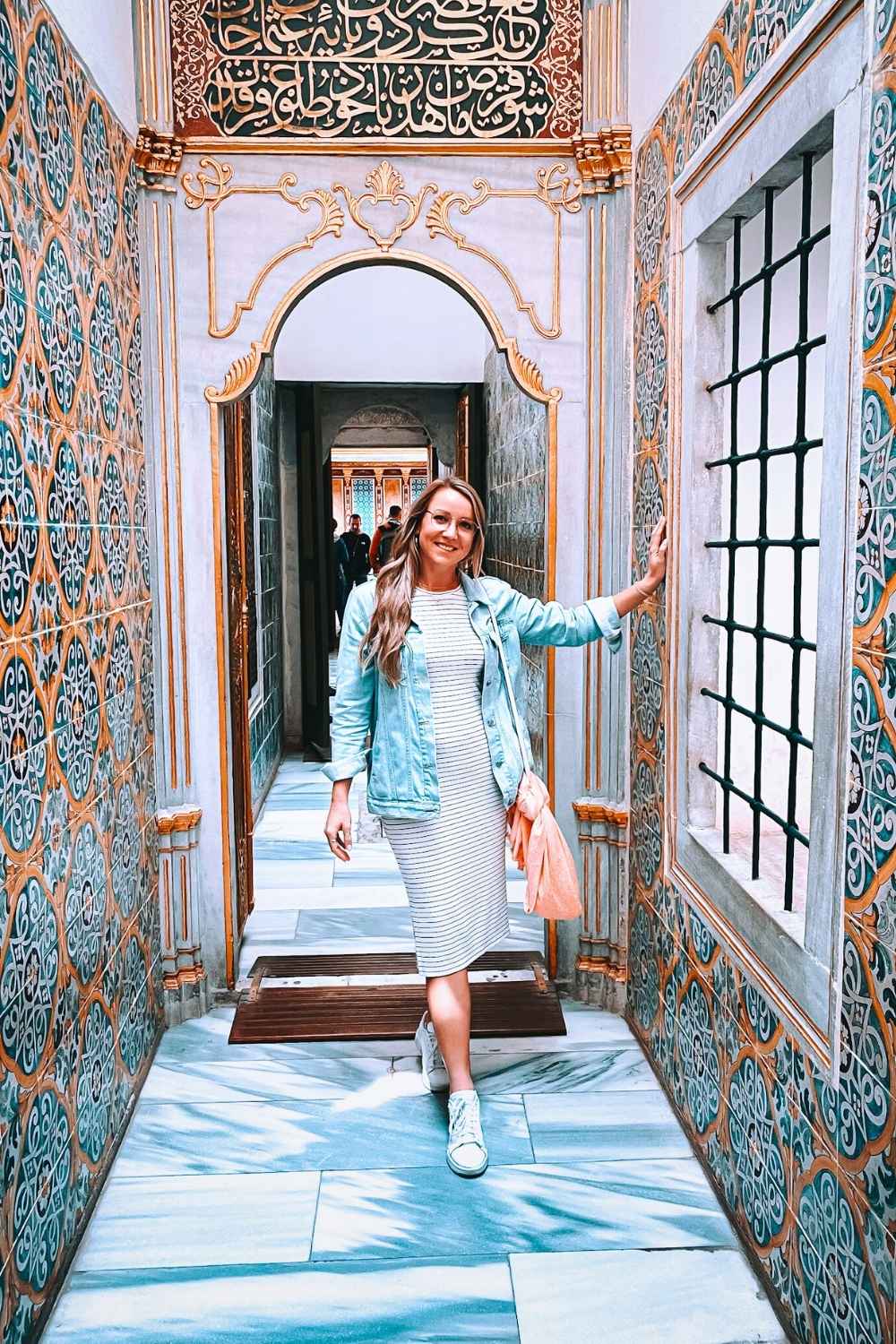 Kate, in a striped dress and denim jacket, poses in a beautifully decorated corridor of Topkapi Palace in Istanbul, surrounded by intricate tile work, golden Arabic calligraphy, and ornate architectural details.