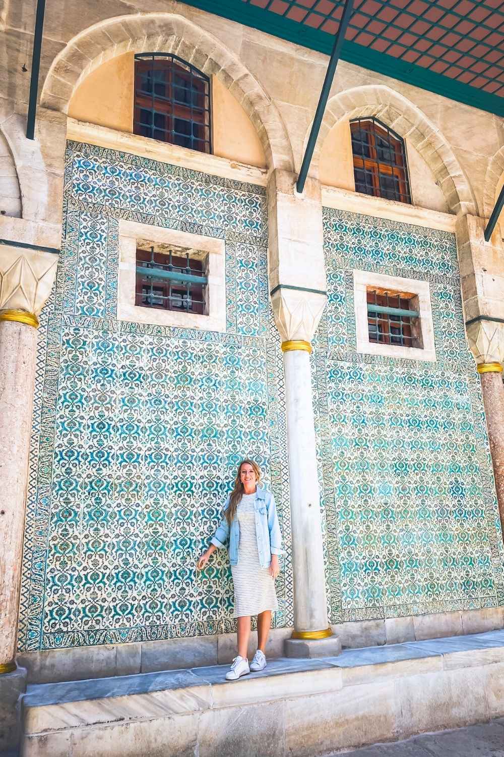 A smiling Kate in a striped dress and denim jacket stands in front of an ornate Iznik tile-covered wall in Topkapi Palace, Istanbul, framed by elegant stone arches and golden-accented columns.