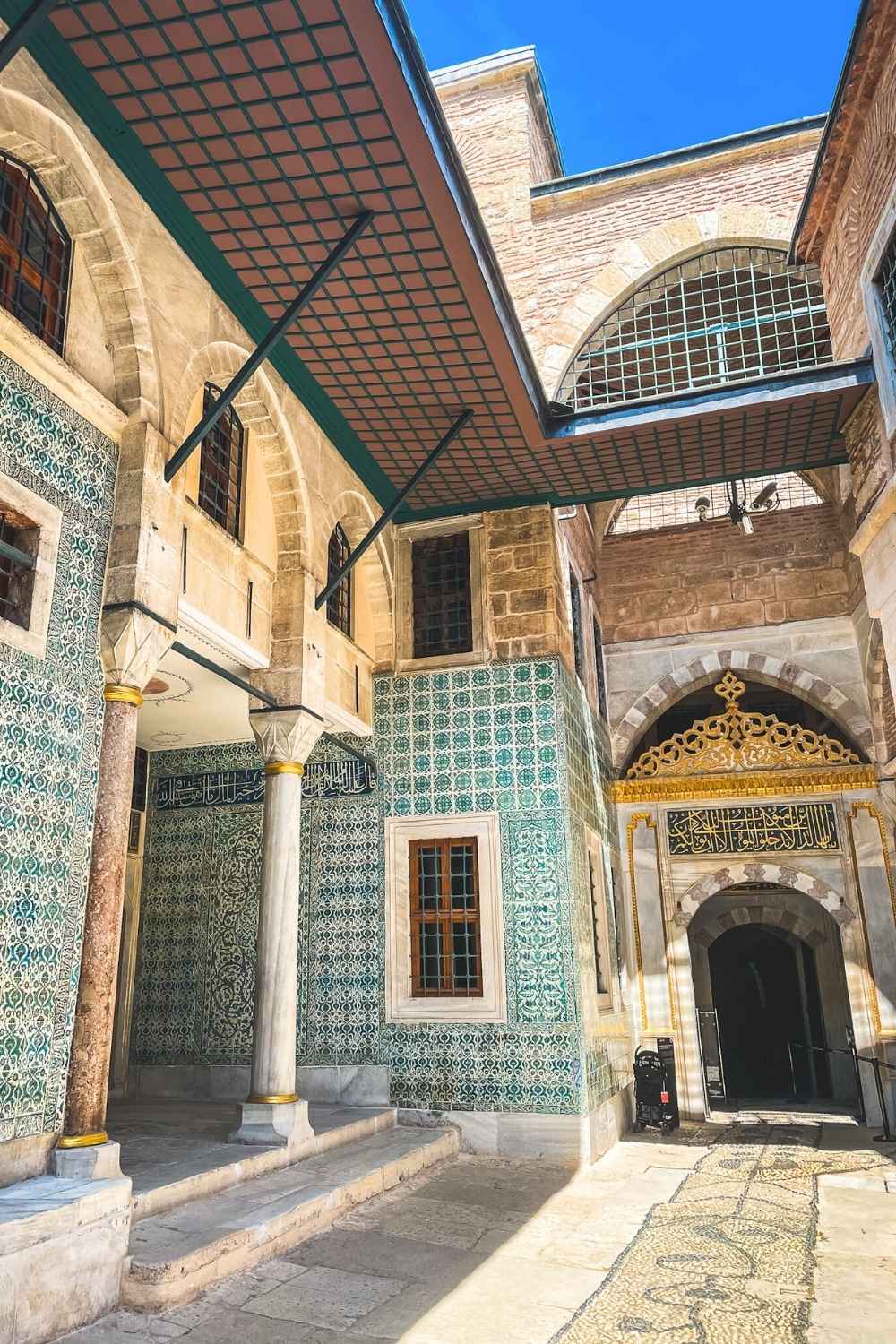 A sunlit courtyard in Topkapi Palace, Istanbul, featuring elegant arches, intricate green and blue Iznik tilework, and a golden Arabic calligraphy inscription above an ornate doorway.