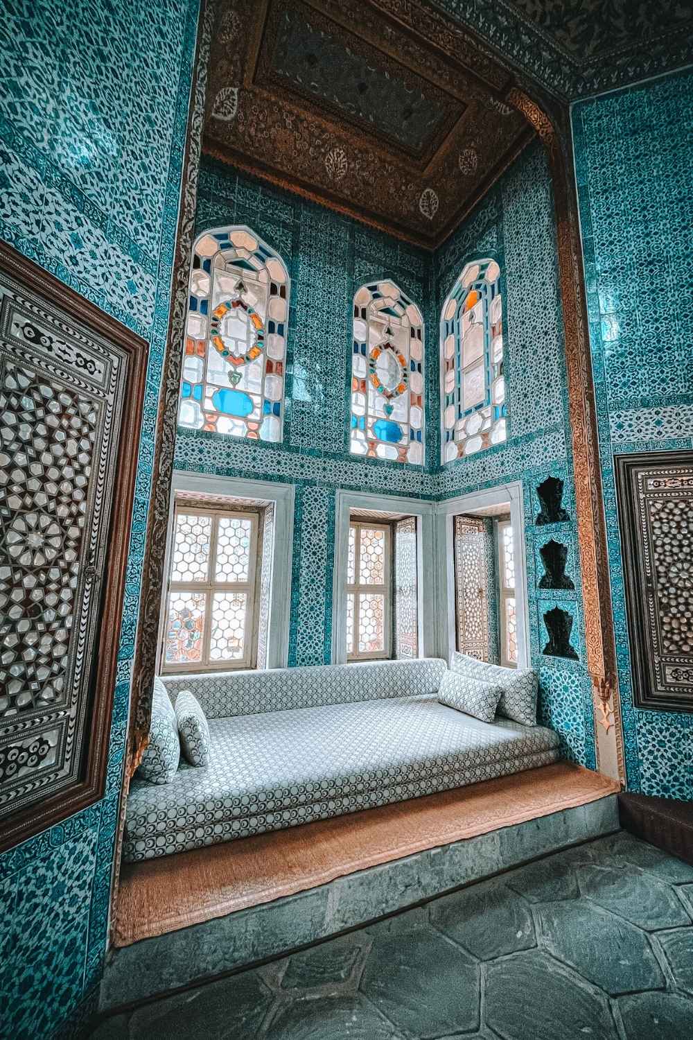 A beautifully decorated room in Topkapi Palace, Istanbul, featuring intricate blue Iznik tilework, ornate stained-glass windows, and a luxurious built-in cushioned seating area with patterned pillows.